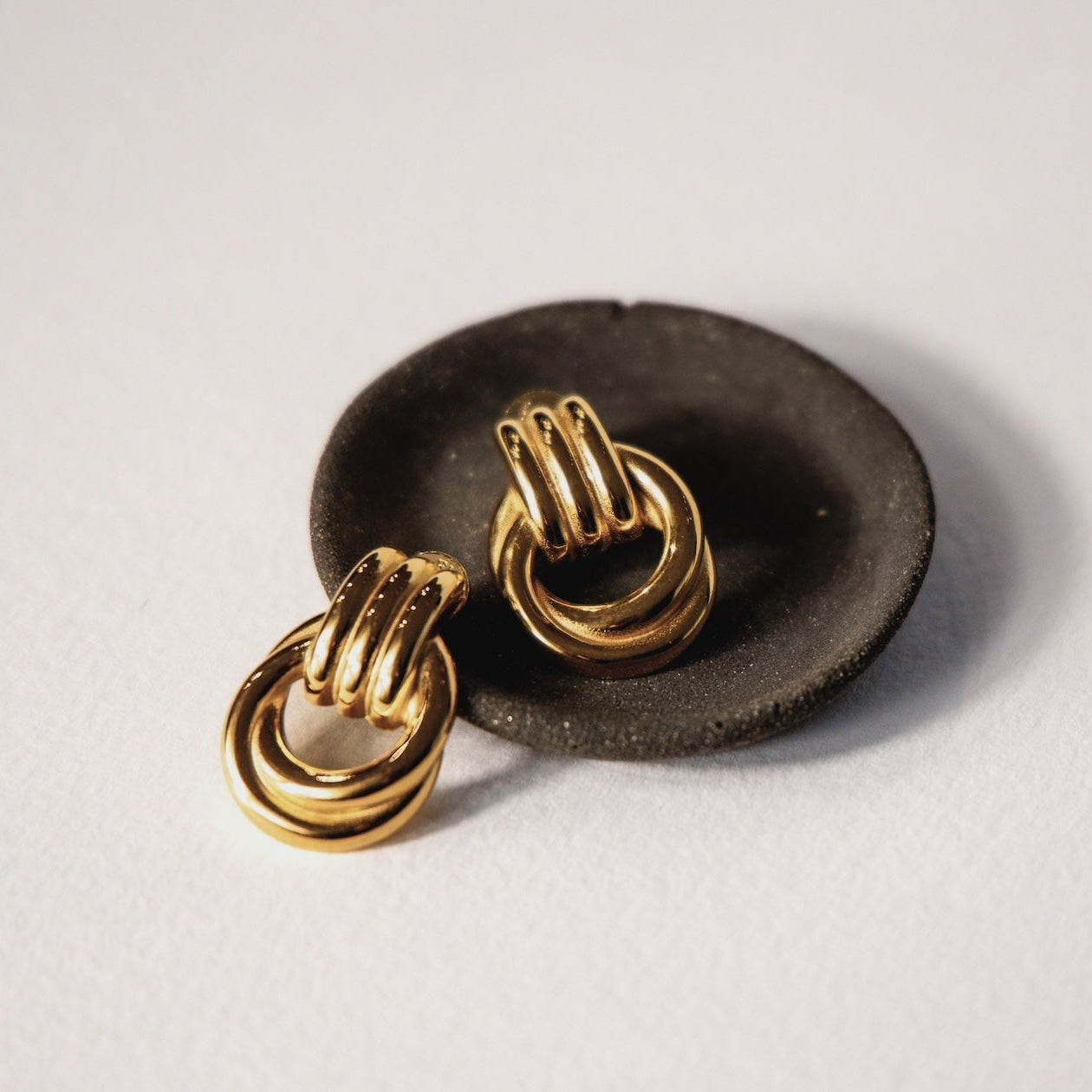 Chunky gold earrings on a black dish against a light gray background