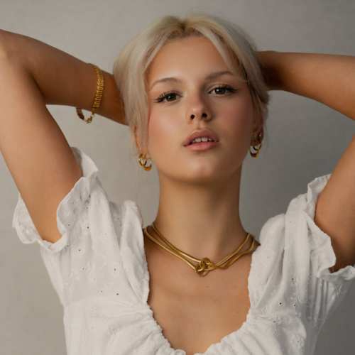 Woman wearing a white dress with gold jewelry against a neutral background
