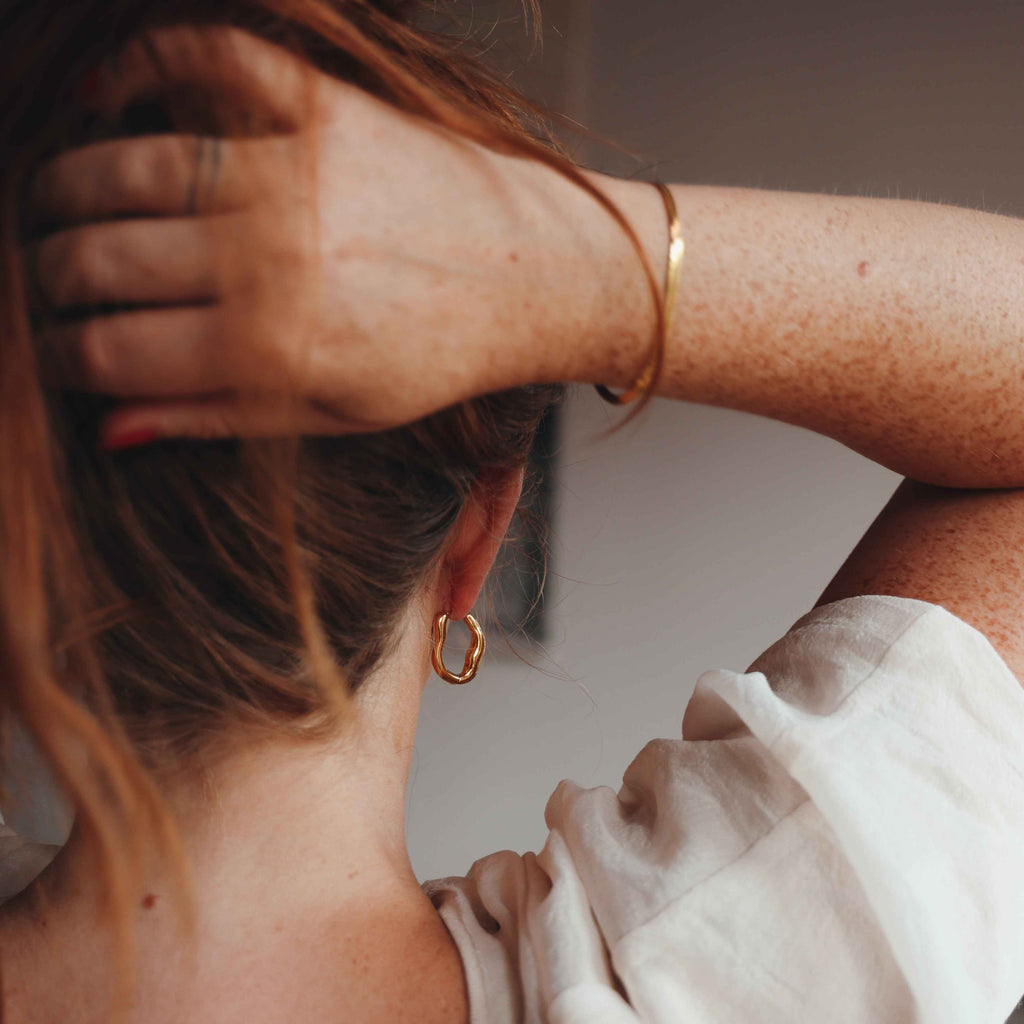 Person with hand in hair wearing a gold earring and bracelet, against a neutral background