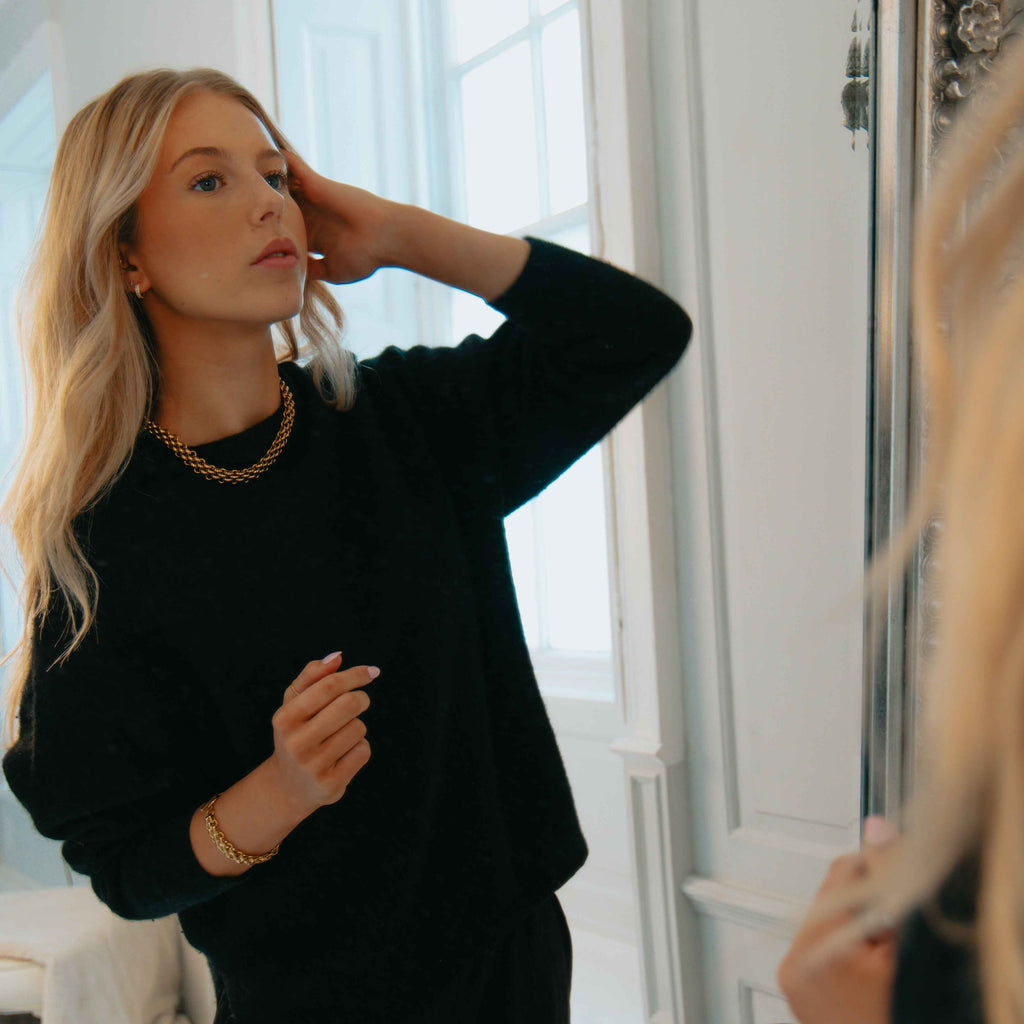 Woman in a chunky gold necklace and black sweater adjusting her hair in front of a mirror.