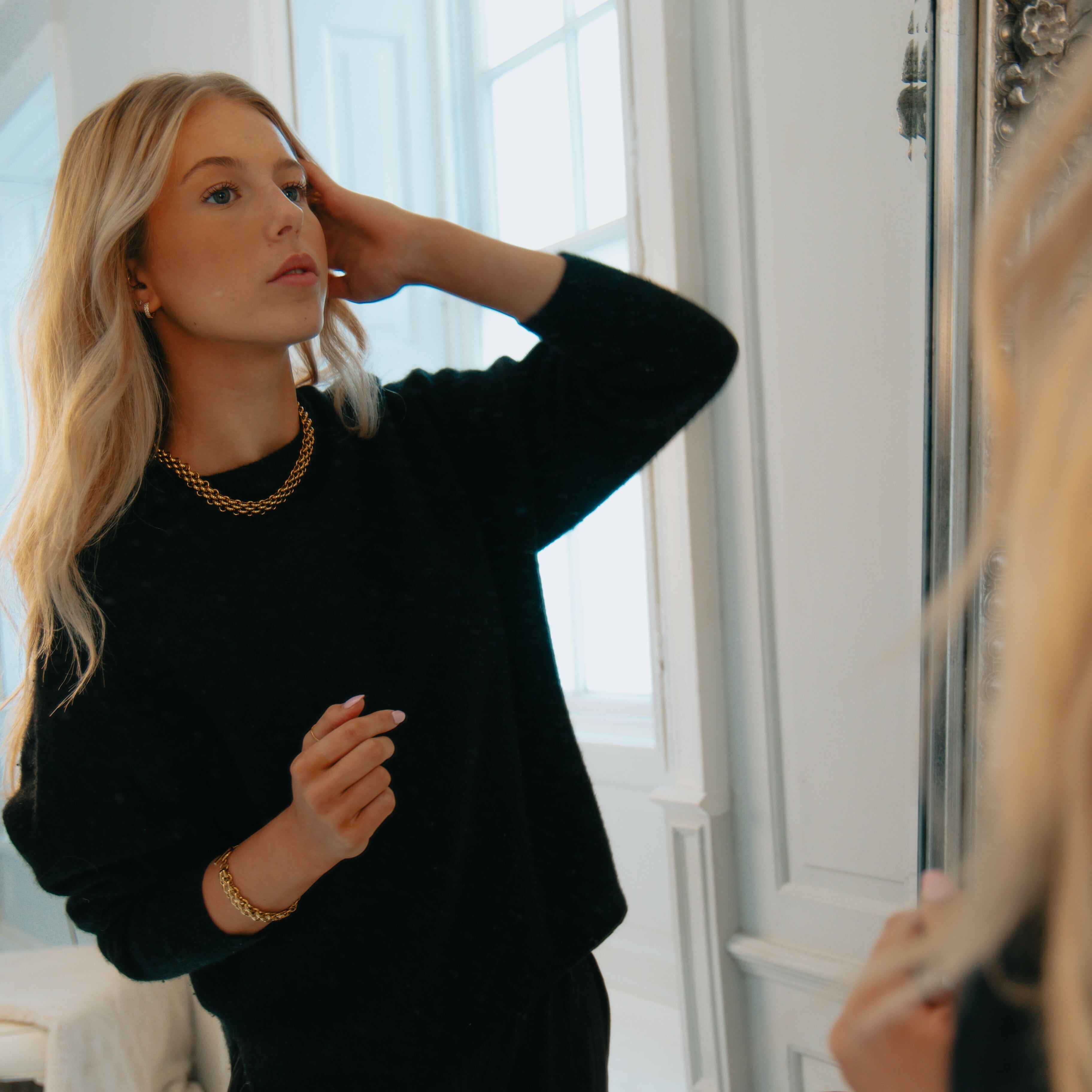 Woman in a chunky gold necklace and black sweater adjusting her hair in front of a mirror.