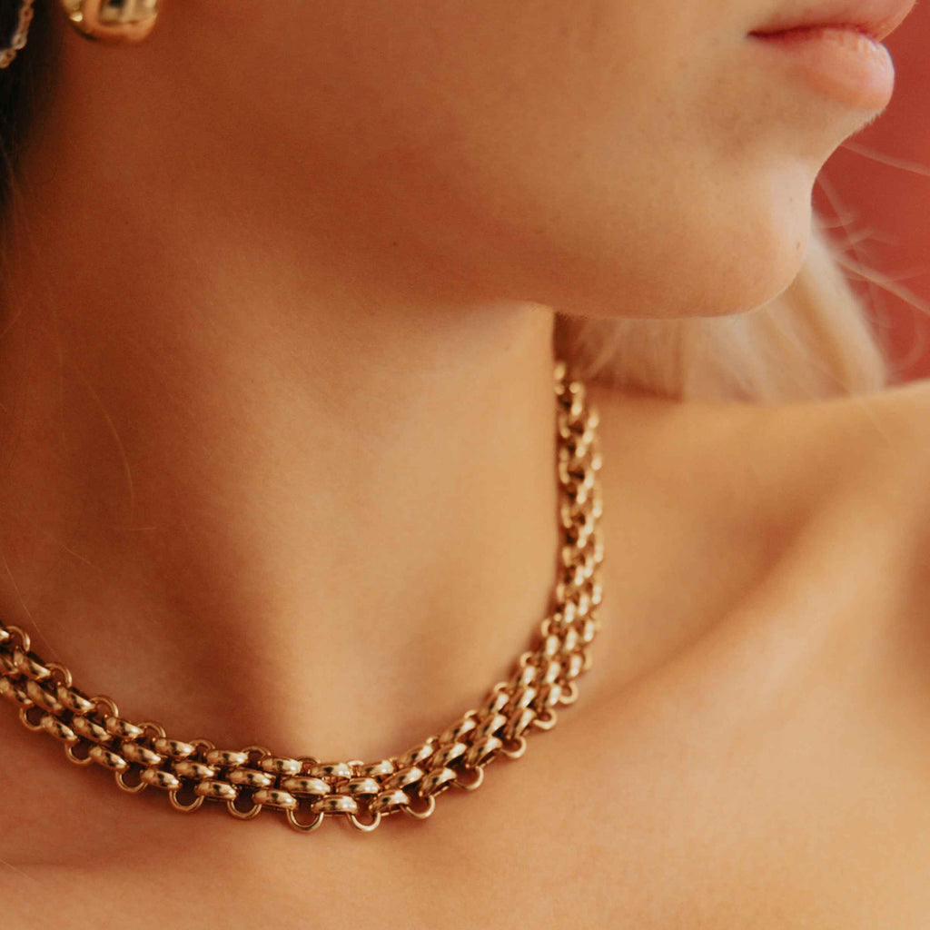 Close-up of a person wearing a gold chain necklace and earrings against a warm-toned background.