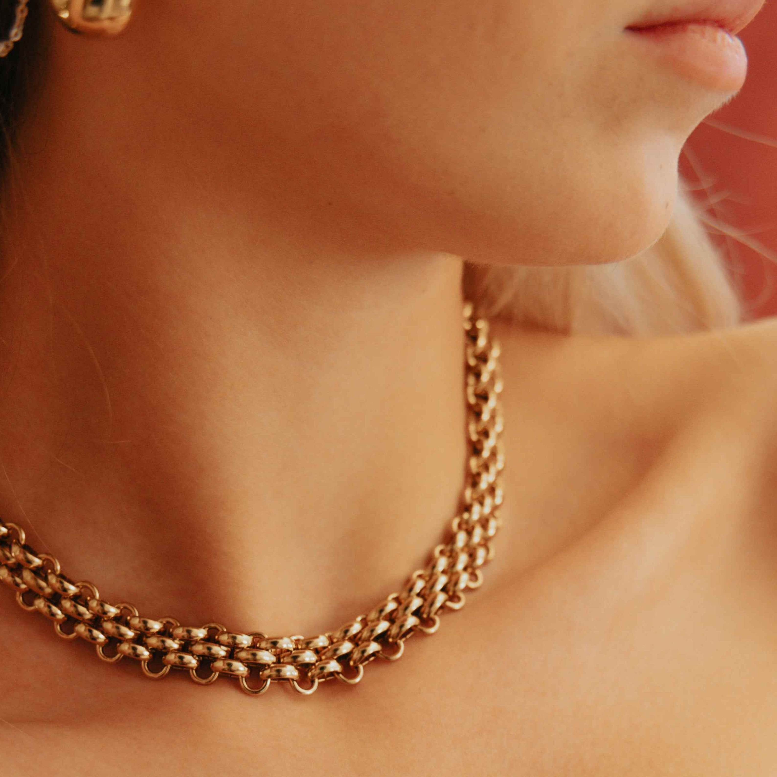 Close-up of a person wearing a gold chain necklace and earrings against a warm-toned background.