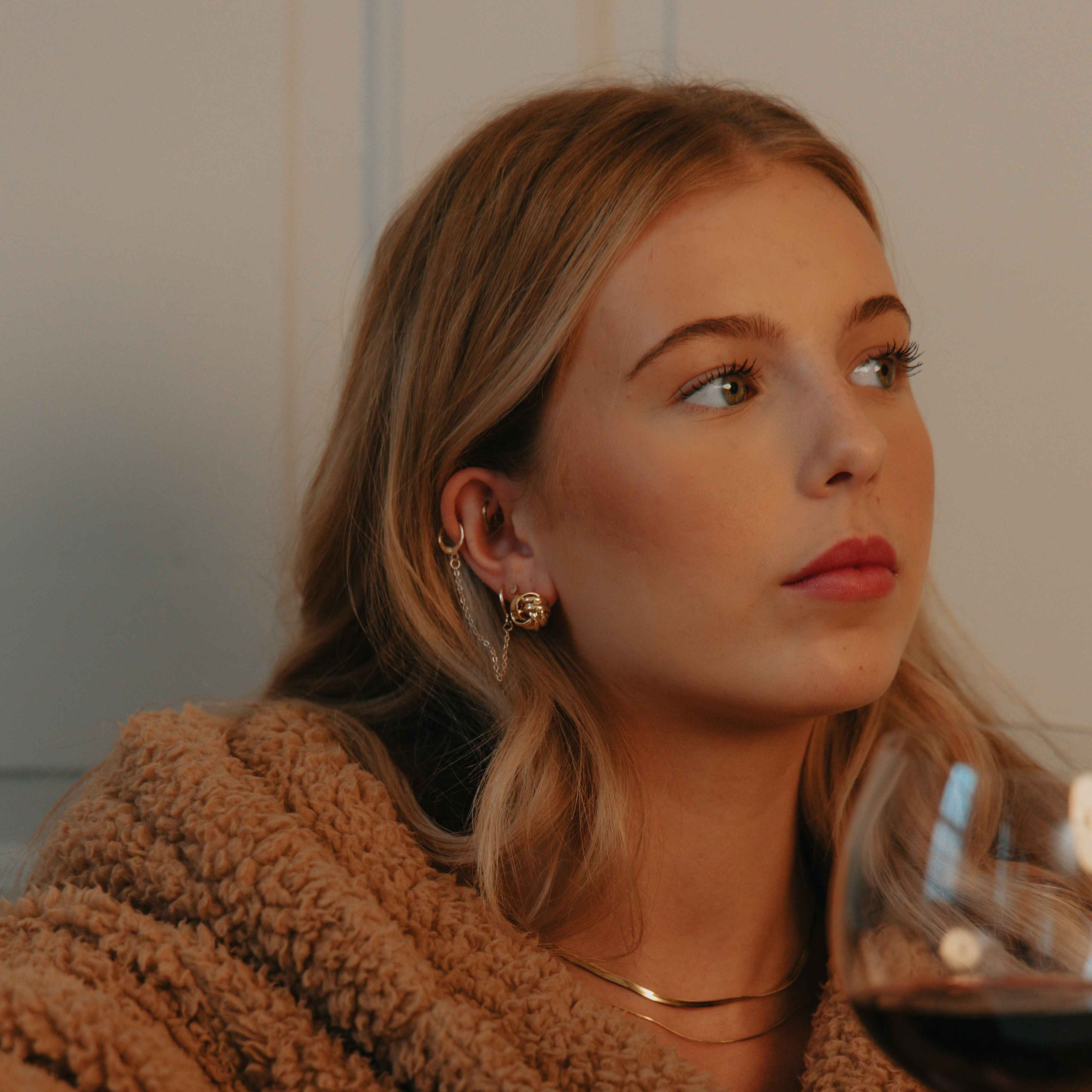 Woman in gold jewellery holding a glass of red wine indoors.