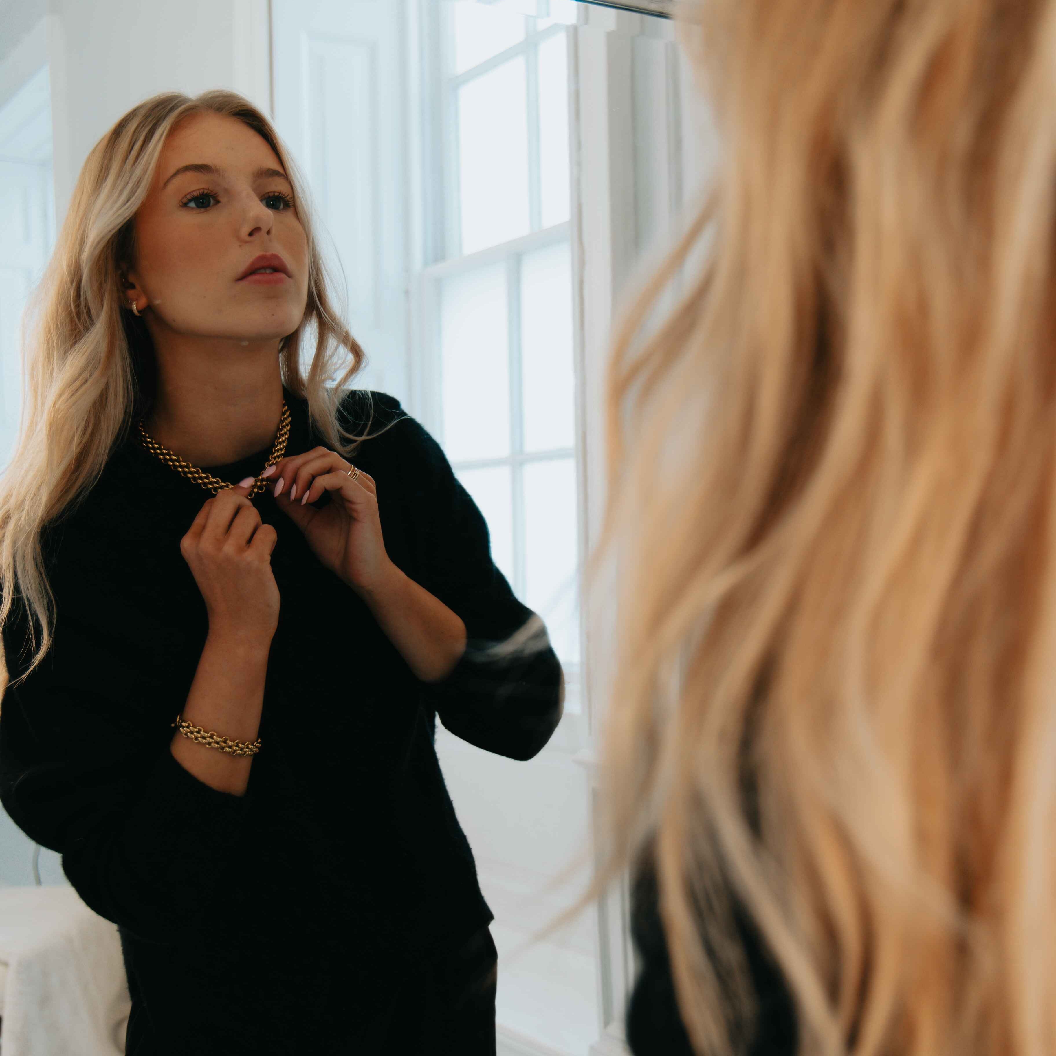 Woman adjusting her gold necklace in front of a large mirror.