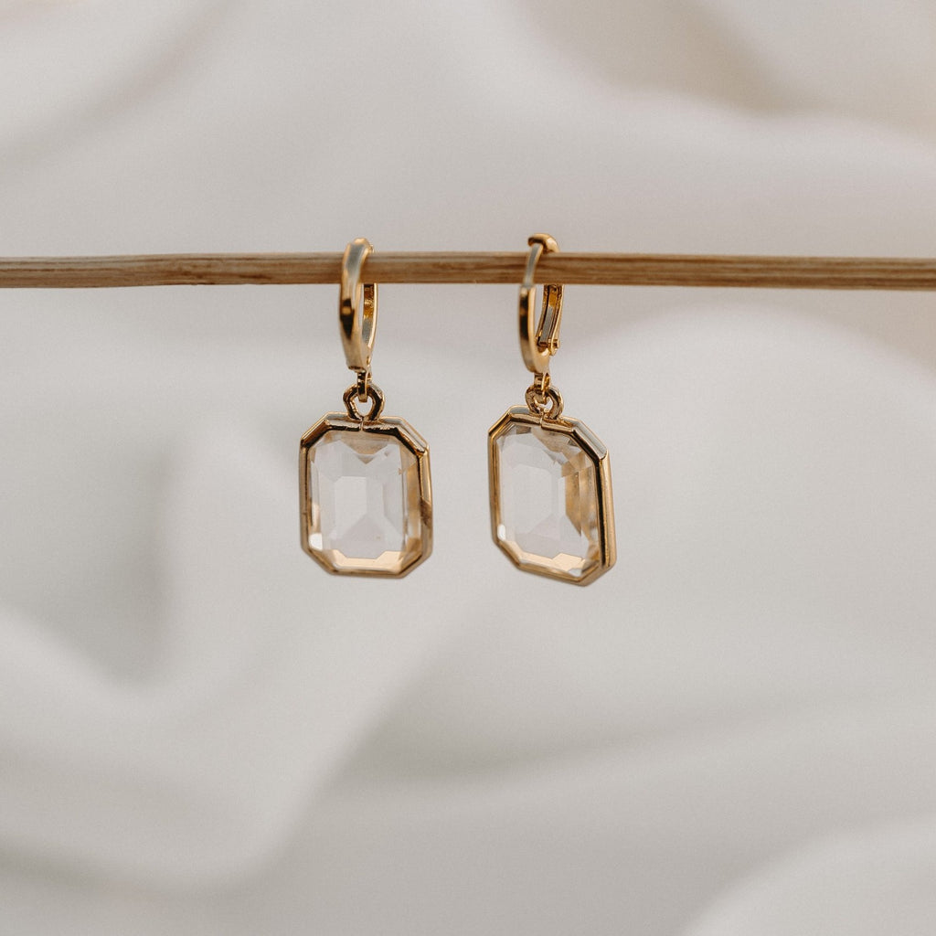 Allana gold huggie earrings with faceted clear drops, hanging from a wooden rail