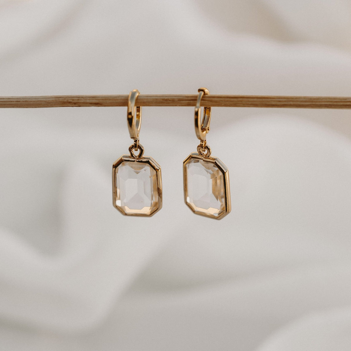 Allana gold huggie earrings with faceted clear drops, hanging from a wooden rail