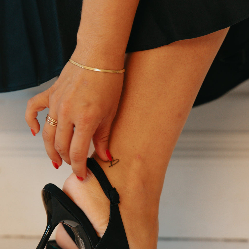 Ava gold herringbone chain bracelet worn on the wrist while adjusting a black slingback heel