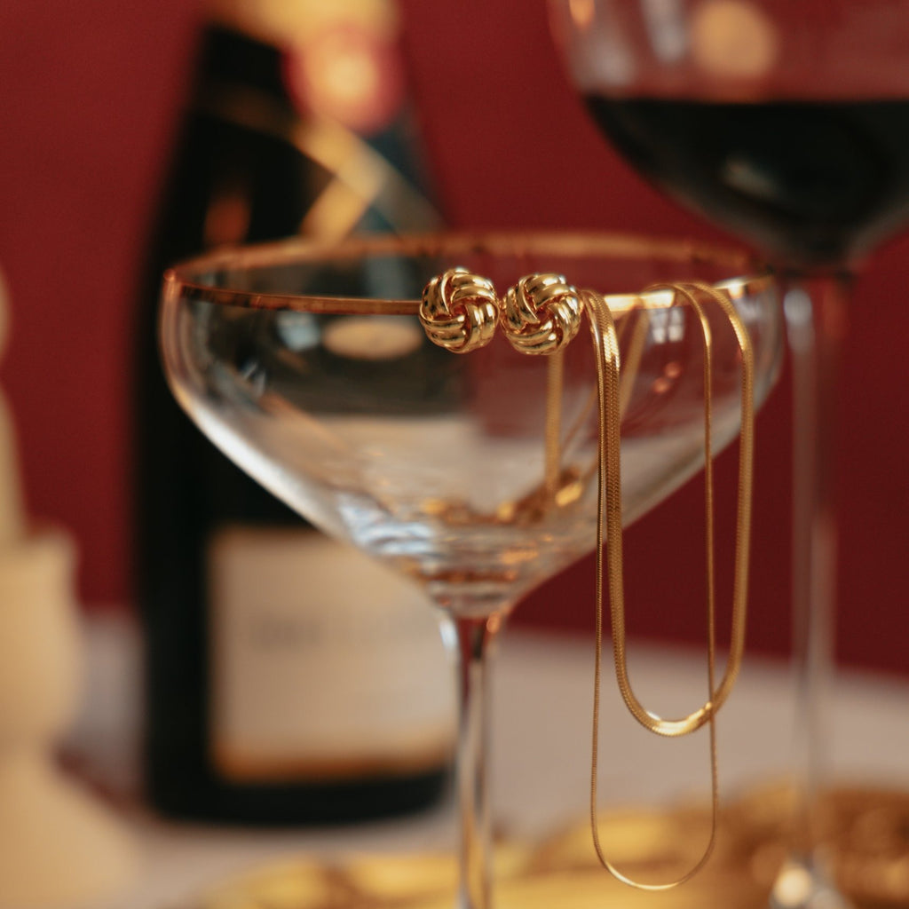 Beatrice knot studs and the Laura double chain necklace resting on the rim of a champagne glass