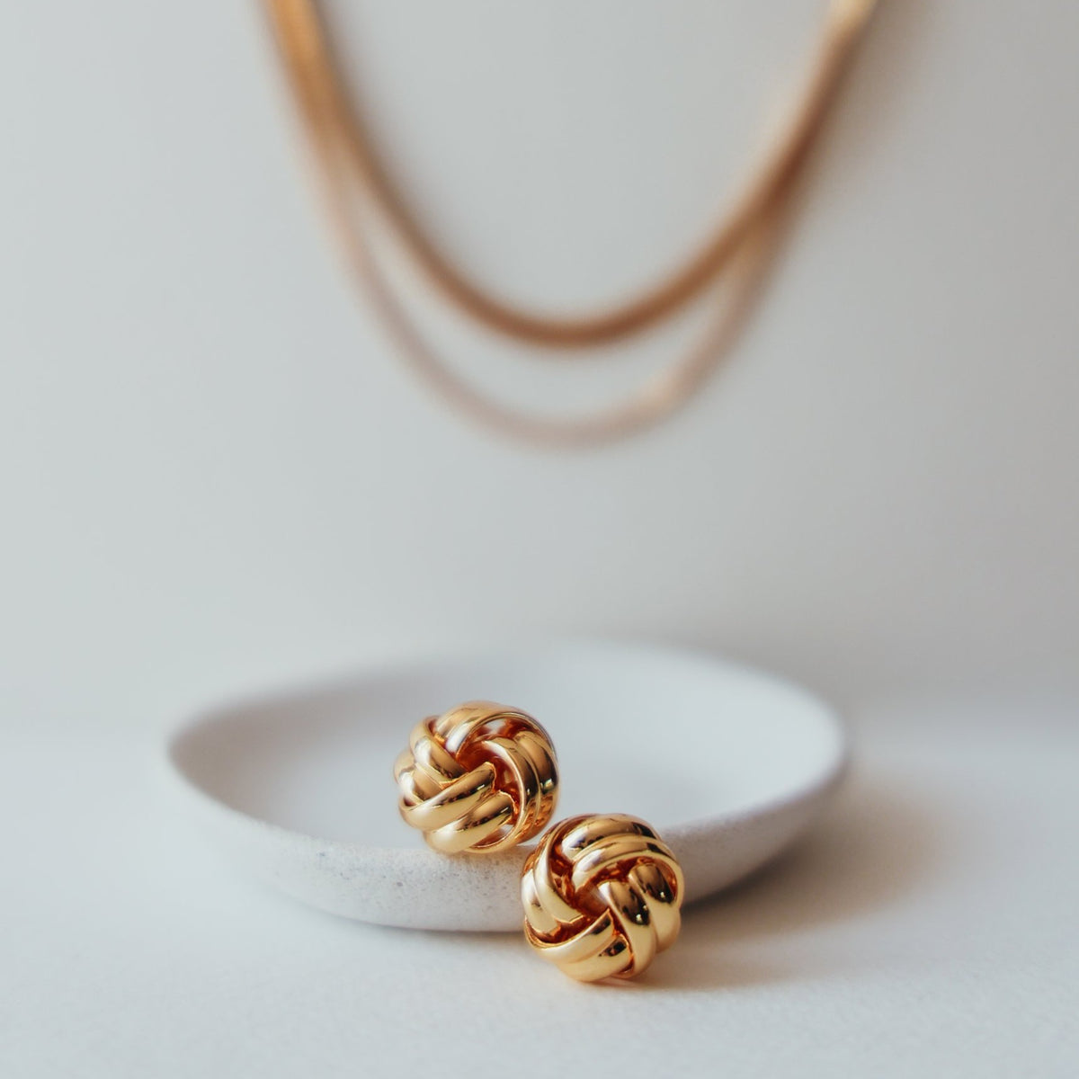Beatrice knot studs on a white dish with the Laura double chain necklace behind