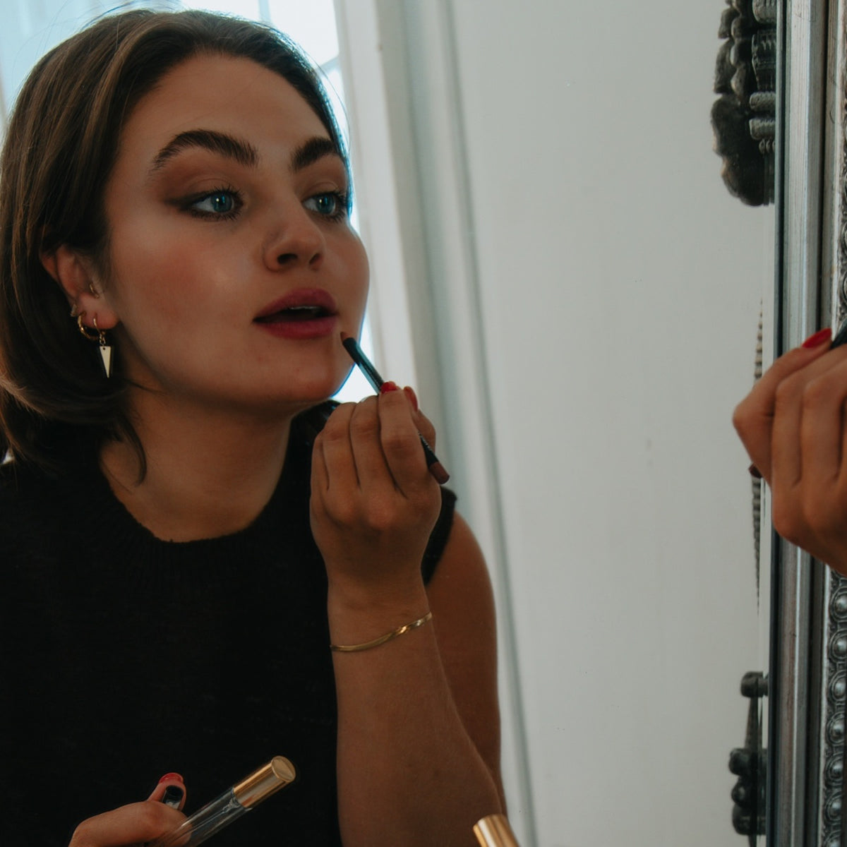 Woman putting on lip liner in a mirror, wearing the Maisie triangle drop huggie earrings