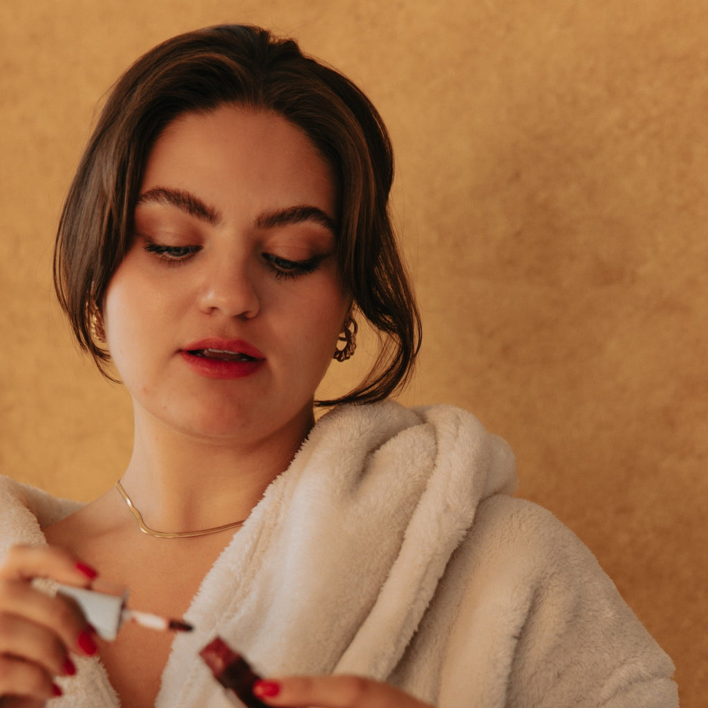 Woman in a cream robe wearing the Sienna gold herringbone chain necklace, putting on lip gloss